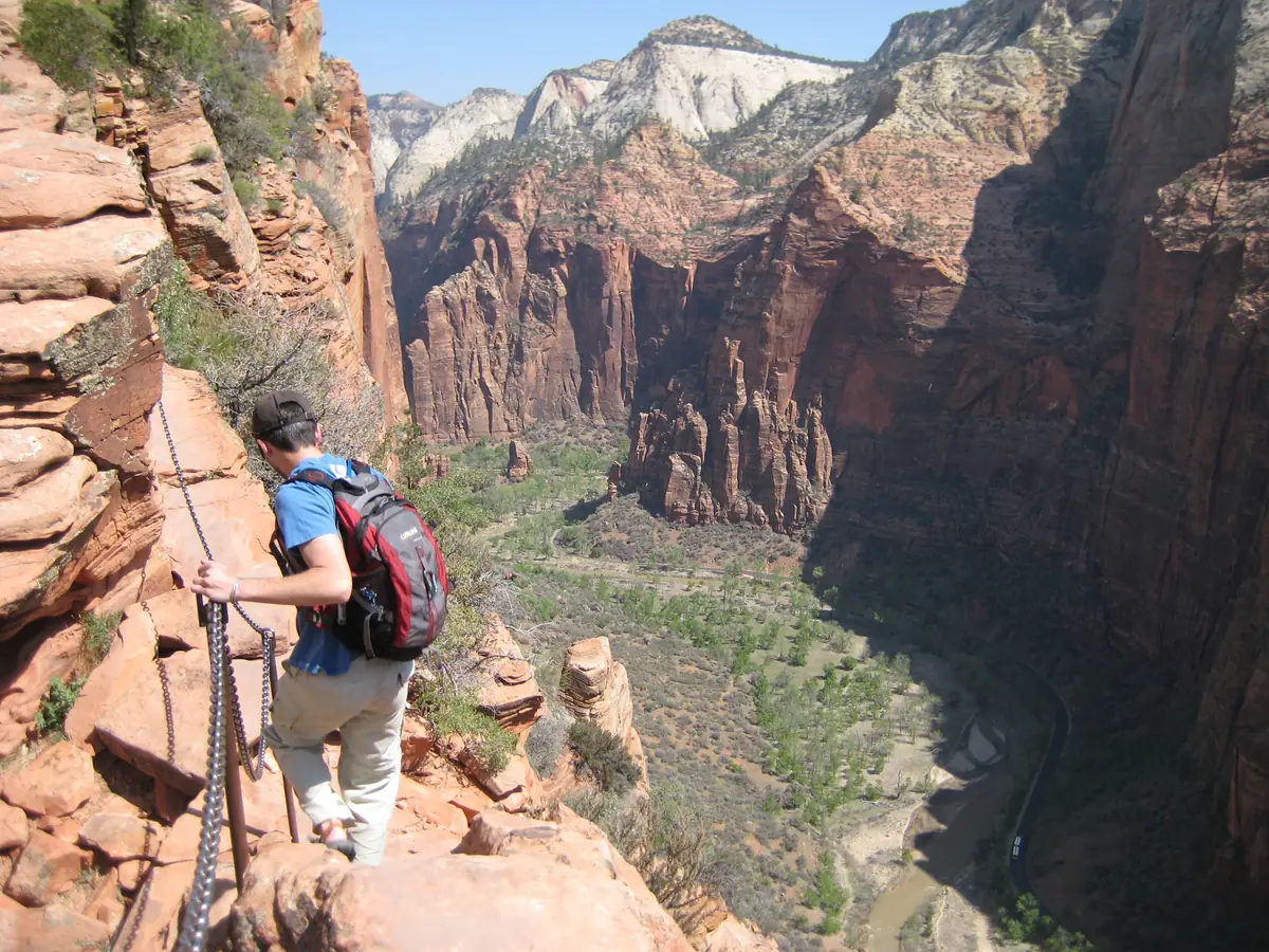 A hiker holding the support chain on the narrow sandstone ridge of the Angels Landing route, with Zion Canyon far below.